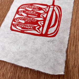 May include: A rectangular piece of textured, off-white paper featuring a red ink block print of three sardines inside an open tin can with a key. The word Sardines is written in pencil on the bottom left, and a signature is on the bottom right.