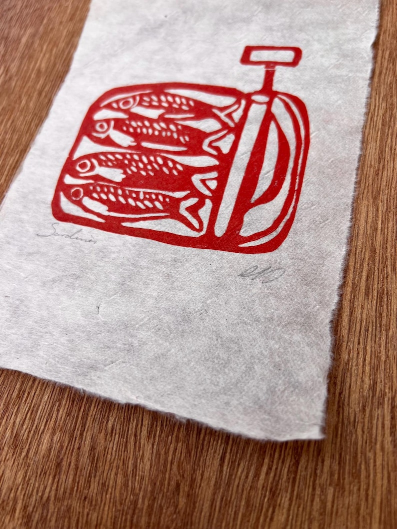May include: A rectangular piece of textured, off-white paper featuring a red ink block print of three sardines inside an open tin can with a key. The word Sardines is written in pencil on the bottom left, and a signature is on the bottom right.
