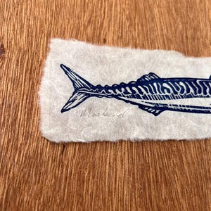 May include: A dark blue ink print of a mackerel fish on a rectangular piece of off-white textured paper with deckled edges. The word Mackerel is handwritten in cursive script in pencil beneath the tail of the fish illustration.
