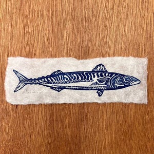 May include: A dark blue ink block print of a mackerel fish on a rectangular piece of textured, off-white handmade paper. The print features detailed line work showing the fish's scales, fins, and eye. The word Mackerel is written in pencil.