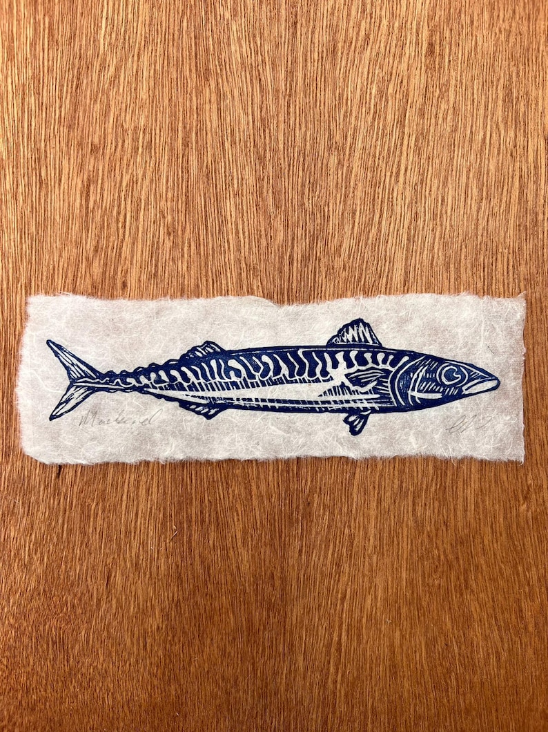 May include: A dark blue ink block print of a mackerel fish on a rectangular piece of textured, off-white handmade paper. The print features detailed line work showing the fish's scales, fins, and eye. The word Mackerel is written in pencil.
