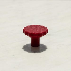 Può includere: Un piccolo pomello per mobili di colore rosso pieno con una parte superiore circolare dal bordo smerlato e una base cilindrica liscia. La superficie superiore presenta una texture fine e sottile che cattura la luce, creando un aspetto rosso opaco.