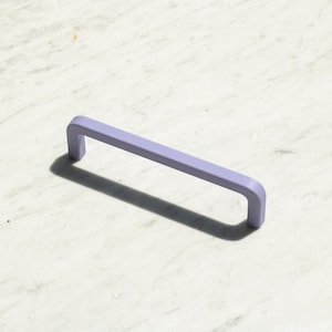 Può includere: Una maniglia per mobili color viola chiaro con una finitura opaca liscia e un design rettangolare a forma di U. La maniglia presenta due punti di fissaggio verticali collegati da una barra orizzontale, creando un profilo pulito e semplice.