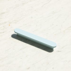 Puede incluir: Un tirador de mueble de color azul claro con una forma rectangular larga y delgada con extremos redondeados. El herraje presenta un acabado mate liso y una superficie superior plana, diseñado para montaje horizontal o vertical en muebles.