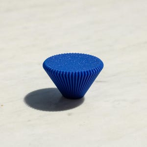 Può includere: Un pomello per mobili di colore blu vibrante con una parte superiore circolare e una base conica scanalata. La superficie superiore presenta una finitura finemente testurizzata, mentre i lati sono definiti da creste verticali.