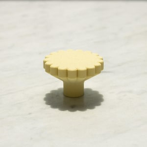 Op de afbeelding: Een lichtgele meubelknop met een cirkelvormige bovenkant met een geschulpte, geribbelde rand en een gladde, cilindrische basis. Het beslag heeft een matte afwerking en een bloemachtige vorm voor een functionele grip op lades of deuren.