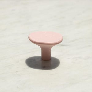 May include: A solid light pink cabinet knob with a smooth, matte finish. The knob features a wide, slightly rounded top surface that tapers down into a cylindrical base, creating a simple, functional shape for furniture hardware.