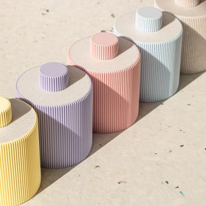 Könnte beinhalten: Fünf zylindrische Behälter mit gerippten Seiten in verschiedenen Pastellfarben: Gelb, Lila, Rosa, Hellblau und Weiß. Die Behälter sind in einer Reihe von links nach rechts angeordnet, wobei der gelbe Behälter vorne und der weiße Behälter hinten ist.