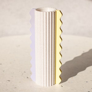 Può includere: Un vaso in ceramica bianca con una texture a coste e un bordo a festoni. Il vaso è decorato con una striscia viola e gialla.