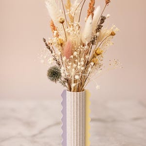 Könnte beinhalten: Ein Trockenblumenarrangement in einer weißen, strukturierten Vase mit violetten und gelben Akzenten. Die Vase hat einen gewellten Rand und ist mit Trockenblumen in Weiß-, Beige- und Brauntönen gefüllt.