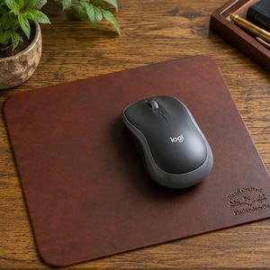 May include: A rectangular dark brown leather mouse pad with rounded corners and a smooth surface. A grey and black wireless computer mouse with the logo logi is positioned on the pad. The text Hand Crafted By FlatlandersCo is embossed.