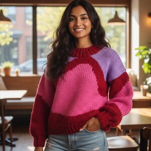 May include: A color-blocked sweater in shades of pink, red, and purple. The sweater has a round neck and long sleeves. The woman is wearing blue jeans.