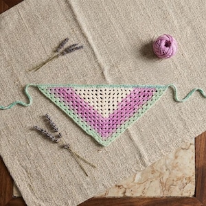 Peut inclure: Un bandana crocheté fait à la main en forme de triangle, avec un dégradé de couleurs allant du blanc au violet, bordé de vert clair. Le bandana est posé sur une surface beige texturée, avec une pelote de fil rose et des brins de lavande.