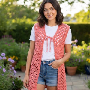 Peut inclure: Un gilet en crochet ajouré de couleur corail avec une fermeture à nouer. Le long gilet est porté sur un t-shirt blanc et un short en jean. Le gilet a un motif rectangulaire et arrive au niveau des genoux.
