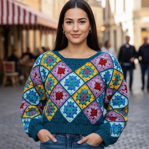 Peut inclure: Une femme porte un pull teal à manches longues avec un motif de crochet géométrique coloré. Le pull présente des carrés bleus, jaunes, rouges et violets. Elle porte un jean bleu et se tient dehors dans une rue pavée.