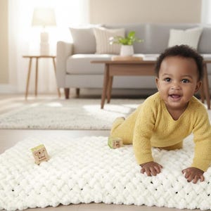 Peut inclure: Un bébé souriant en tenue jaune rampe sur une couverture blanche épaisse et tricotée. Deux blocs de bois avec des motifs imprimés sont sur la couverture. Un canapé, une table basse et une lampe sont en arrière-plan.