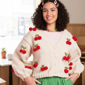 May include: A cream-coloured cardigan with red cherry appliqués and green leaves. The button-down sweater has a V-neck and long sleeves. The model is wearing a pink top and a green skirt.