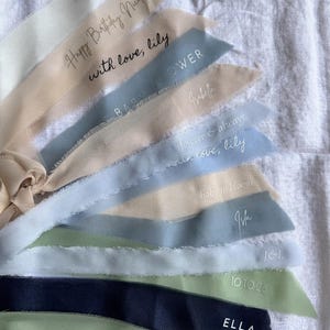 May include: A collection of frayed-edge fabric ribbons in shades of cream, beige, blue, sage green, and navy. Each ribbon features custom printed text including names like Isla, Isabella, Ella, and phrases such as Happy Birthday, with love, lily, and 10 10 26.