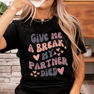 May include: Black t-shirt with the text "Give me a break, my partner died" in pink and brown lettering with hearts.