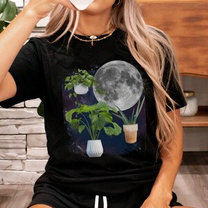 May include: A black t-shirt with a graphic of three potted plants in front of a full moon. The plants are a monstera, a snake plant, and a succulent. The moon is white and the background is a dark blue.