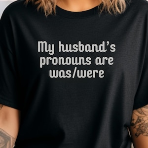 May include: Black t-shirt with the text "My husband's pronouns are was/were" in white. The shirt is a crew neck and has short sleeves. The text is centered on the front of the shirt.