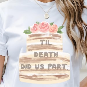 May include: White t-shirt featuring a two-tiered cake design with pink roses and the text "Til Death Did Us Part." The cake has a watercolor style with brown and beige tones. The shirt is a casual, everyday wear item.