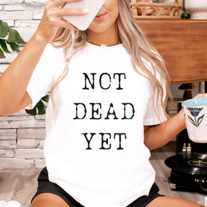 May include: A white t-shirt with the text "NOT DEAD YET" printed on it. The shirt is being worn by a person sitting on a rug with a record player and a mug in the background.