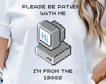 May include: A white short-sleeved t-shirt featuring a pixel art graphic of a vintage computer monitor displaying the text Hi. Above and below the graphic, the black pixelated text reads PLEASE BE PATIENT WITH ME and I'M FROM THE 1900S.