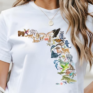 May include: White t-shirt with a colorful illustration of the state of Florida made up of various animals, including a bear, a raccoon, a panther, a dolphin, a manatee, a turtle, and a crocodile.
