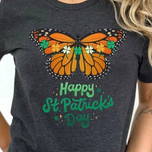 May include: A dark gray t-shirt with a graphic of an orange butterfly with green shamrocks. The text "Happy St. Patrick's Day" is printed below the butterfly.