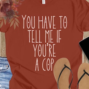 May include: A rust-colored t-shirt with the text "You have to tell me if you're a cop" printed in white.