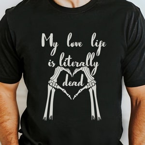 May include: Black t-shirt with the text "My love life is literally dead" in a white, stylized font. The design includes a heart shape formed by two white skeleton hands. The shirt is a casual wear item.