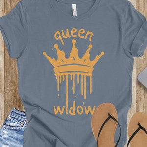 May include: A slate blue t-shirt with the words "queen widow" and a dripping gold crown graphic. The shirt is paired with denim shorts and brown flip-flops. A smartphone and key are also visible.