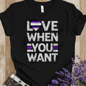 Könnte beinhalten: Ein schwarzes Kurzarm-T-Shirt mit dem weißen, abgenutzten Schriftzug LOVE WHEN YOU WANT. Der Buchstabe O ist durch ein Herz mit horizontalen Streifen in Weiß, Grau, Schwarz und Lila ersetzt. Eine lila Linie akzentuiert das Wort YOU.