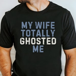 May include: A black t-shirt with the text "MY WIFE TOTALLY GHOSTED ME" in a distressed blue font.