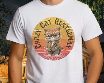 May include: A white t-shirt featuring a graphic of a cat wearing a tan suit, a brown tie, and black glasses. The cat is centered within a circular sunset-colored background with the text CRAZY CAT GENTLEMAN arched around the top of the circle.