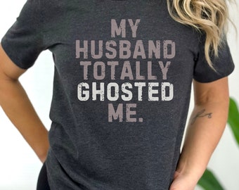 May include: A dark gray t-shirt with the text "MY HUSBAND TOTALLY GHOSTED ME." printed in white and light pink.