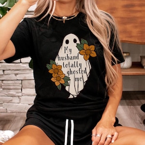 May include: Black t-shirt with a white ghost graphic and the text "My husband totally ghosted me". The ghost has two eyes and a mouth. There are orange flowers around the ghost.