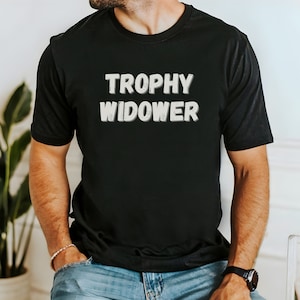 May include: Black crew neck t-shirt with the text "TROPHY WIDOWER" in a bold, 3D-style font. The text is in white with a subtle shadow effect. The shirt is a casual style, suitable for everyday wear.