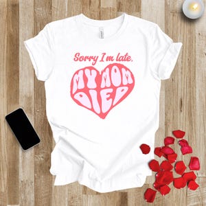 May include: White t-shirt with the text "Sorry I'm late. MY MOM DIED" in pink, inside a pink heart. A black smartphone and red rose petals are on a wooden surface. The t-shirt is the main focus.