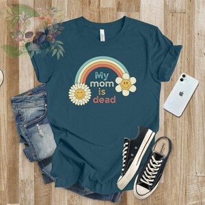 May include: A teal blue t-shirt with a rainbow and two yellow flowers with smiley faces. The text on the shirt reads "My mom is dead".