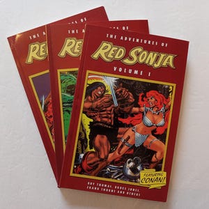 Może przedstawiać: Stos czterech czerwonych komiksów z przygodami Red Sonji. Okładka górnego komiksu przedstawia Red Sonję w czerwonym bikini, trzymającą miecz i stojącą obok muskularnego mężczyzny. Tekst na okładce brzmi "The Adventures of Red Sonja, Volume I, Featuring Conan!"