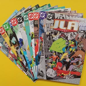 Op de afbeelding: Een verzameling DC Comics-strips uitgespreid tegen een gele achtergrond. De strips tonen verschillende covers met het DC-logo en superheldenillustraties. De JLA Year One-strip is zichtbaar.