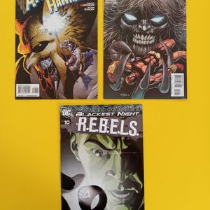 以下が含まれることがあります： 「Blackest Night」というタイトルの3冊のコミックが展示されています。表紙には、スーパーヒーローと悪役の鮮やかなイラストが描かれています。1冊のコミックは「The Atom and Hawkman」、もう1冊は「Outsiders」、3冊目は「R.E.B.E.L.S.」です。
