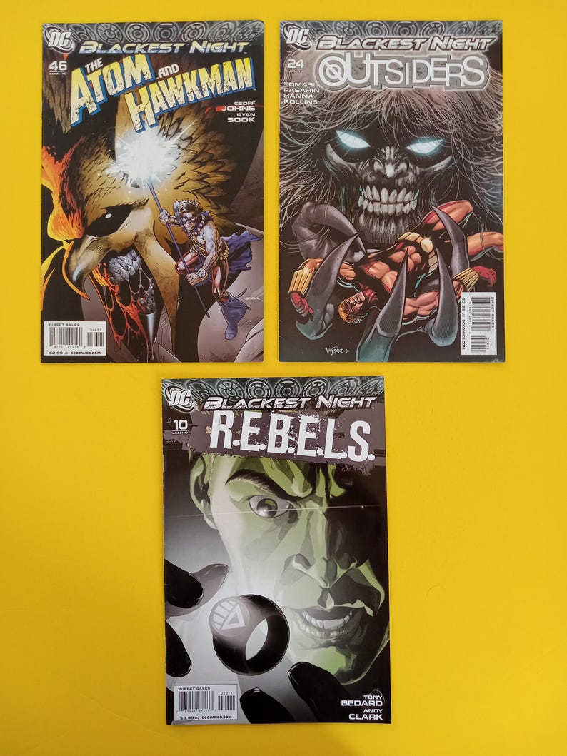 以下が含まれることがあります： 「Blackest Night」というタイトルの3冊のコミックが展示されています。表紙には、スーパーヒーローと悪役の鮮やかなイラストが描かれています。1冊のコミックは「The Atom and Hawkman」、もう1冊は「Outsiders」、3冊目は「R.E.B.E.L.S.」です。