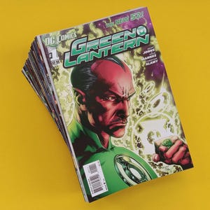 May include: A stack of comic books featuring the Green Lantern issue number 1 from DC Comics The New 52 series. The cover displays a portrait of a character with red skin, a green uniform, and a glowing green ring on a finger against a green background.
