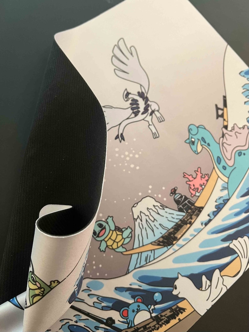 Op de afbeelding: Een witte muismat met een Japans golfontwerp met verschillende Pok&eacute;mon-personages, waaronder Squirtle, Bulbasaur en een wit kat-achtig Pok&eacute;mon. De muismat heeft een zwarte, stoffen achterkant.