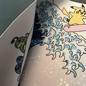 Op de afbeelding: Een witte muismat met een blauw en wit golfontwerp. Het golfontwerp doet denken aan de Grote Golf voor Kanagawa. Er zijn cartoonfiguren op de muismat, waaronder een gele Pikachu die op een golf rijdt en een groene rups.
