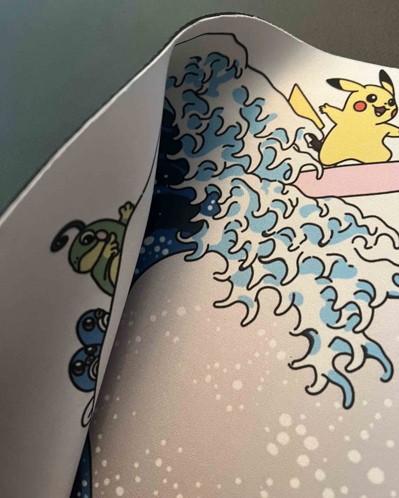 Op de afbeelding: Een witte muismat met een blauw en wit golfontwerp. Het golfontwerp doet denken aan de Grote Golf voor Kanagawa. Er zijn cartoonfiguren op de muismat, waaronder een gele Pikachu die op een golf rijdt en een groene rups.