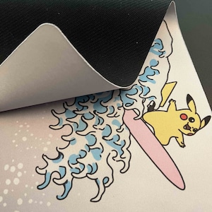 Op de afbeelding: Een witte muismat met een cartoon afbeelding van Pikachu die op een roze surfplank op een blauwe golf rijdt. De muismat heeft een zwarte rubberen achterkant.